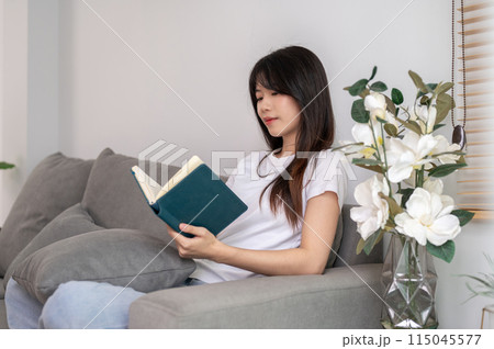 A young, relaxed Asian woman is reading a book on the sofa in the living room at home. 115045577