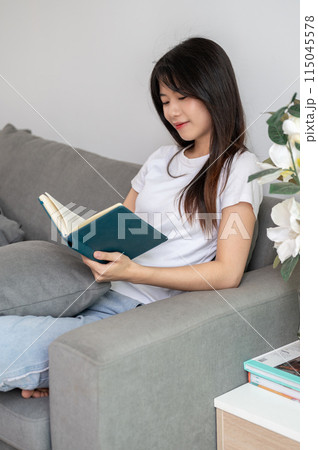 A young, relaxed Asian woman is reading a book on the sofa in the living room at home. A young, relaxed Asian woman is reading a book on the sofa in the living room at home. 115045578