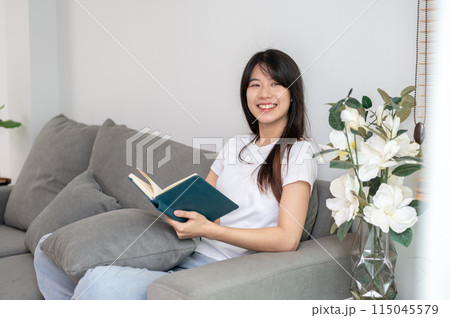 A beautiful young Asian woman is daydreaming while reading a book on the sofa in the living room. A beautiful young Asian woman is daydreaming while reading a book on the sofa in the living room. 115045579