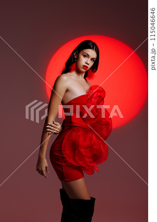 A young girl in a red dress posing in the studio against the backdrop of a red spotlight. A young girl in a red dress posing in the studio against the backdrop of a red spotlight. 115046646