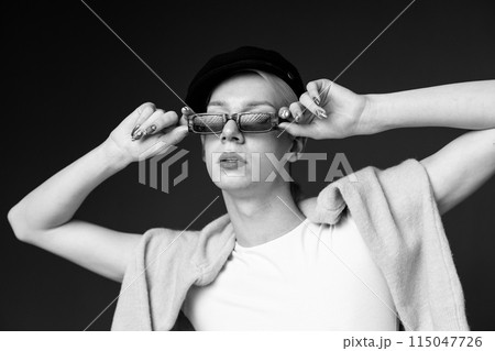 Black and white photo of young man with make up on his face wearing sunglasses. Man wearing fashionable sweater and cap. Man posing for a photo. 115047726