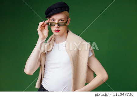 Young blond man with make up on his face and sunglasses. Man wearing fashionable white t-shirt and black cap. Man isolated on green background. 115047736