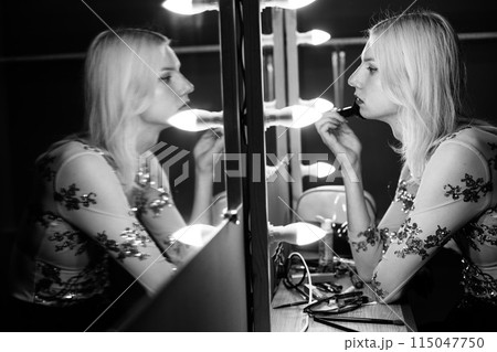 Young man holding brushes and doing make up on his face. Man wearing fashionable t-shirt. Man looking in a studio mirror. 115047750