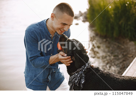 Handsome young male in casual outfit playing with cute dog while standing near the lake. Boy wearing blue shirt and white jeans shorts. Dog has a white and black scarf on his neck. Handsome young male in casual outfit playing with cute dog while standing near the lake. Boy wearing blue shirt and white jeans shorts. Dog has a white and black scarf on his neck. 115047849