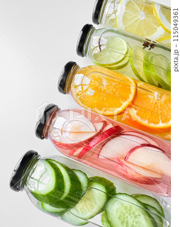 Stacked glass bottles with detox drinks with cucumber, apple, orange, and lemon slices on white background. Vertical orientation, refreshing beverage concept for design and print. Stacked glass bottles with detox drinks with cucumber, apple, orange, and lemon slices on white background. Vertical orientation, refreshing beverage concept for design and print. 115049361