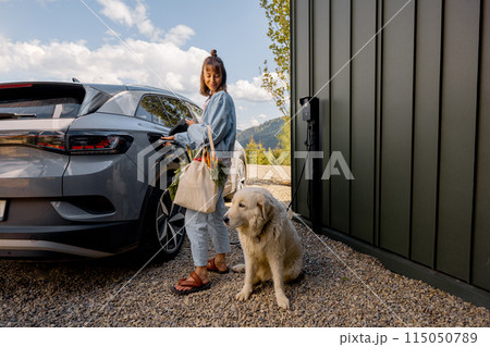 Woman plugging a charger into electric vehicle, with a dog near house 115050789