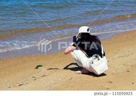 海辺、砂浜でしゃがんで携帯を持ちながら海を眺める一人の女性　長い黒髪に白いキャップ 115050925