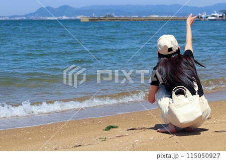海辺、砂浜でしゃがんで海を眺める一人の女性　長い黒髪に白いキャップ 115050927