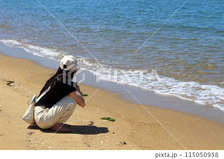 海辺、砂浜でしゃがんで海を眺める一人の女性 長い黒髪に白いキャップ 海辺、砂浜でしゃがんで海を眺める一人の女性 長い黒髪に白いキャップ 115050938