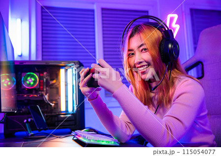 Smiling Asian woman live stream she play video game via smartphone at home neon lights living room, Gamer playing online game application on mobile phone wear gaming headphones looking to camera 115054077