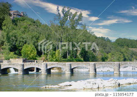Serene Stone Bridge Over Tranquil River in Lush Forest Landscape with Blue Sky and Clouds 115054177