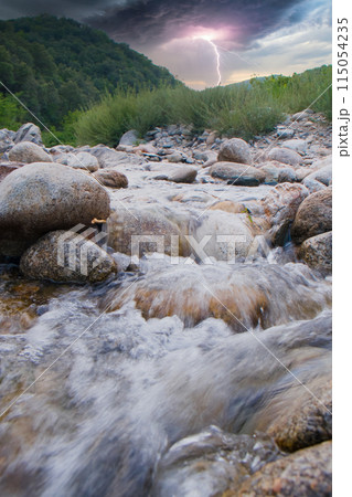 River stream view during lightning storm, with mountains and lush forest in background River stream view during lightning storm, with mountains and lush forest in background 115054235