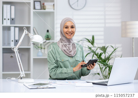 Portrait of successful and happy businesswoman financier, woman in hijab smiling and looking at camera, using calculator at work, accountant working with papers using laptop. Portrait of successful and happy businesswoman financier, woman in hijab smiling and looking at camera, using calculator at work, accountant working with papers using laptop. 115057237