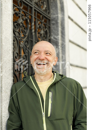 Content bald senior man with beard wearing a green Marmot jacket, smiling in a sunlit urban setting Content bald senior man with beard wearing a green Marmot jacket, smiling in a sunlit urban setting 115057966