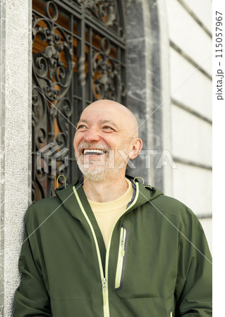 Content bald senior man with beard wearing a green Marmot jacket, smiling in a sunlit urban setting 115057967