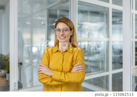 Confident businesswoman in a yellow shirt and glasses standing with arms crossed in a modern office setting. Confident businesswoman in a yellow shirt and glasses standing with arms crossed in a modern office setting. 115062513