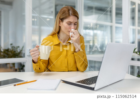 A woman in a yellow shirt sits at her desk, drinking tea and blowing her nose, indicating illness and discomfort at work. A woman in a yellow shirt sits at her desk, drinking tea and blowing her nose, indicating illness and discomfort at work. 115062514