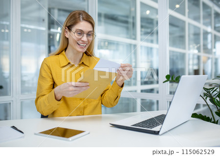 Happy woman smiling and reading a letter at her desk in a modern office. She appears pleased with the contents. Happy woman smiling and reading a letter at her desk in a modern office. She appears pleased with the contents. 115062529