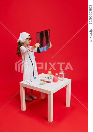 Girl dressed as doctor examines X-ray, standing by table with medical supplies against vibrant red background. Girl dressed as doctor examines X-ray, standing by table with medical supplies against vibrant red background. 115062902