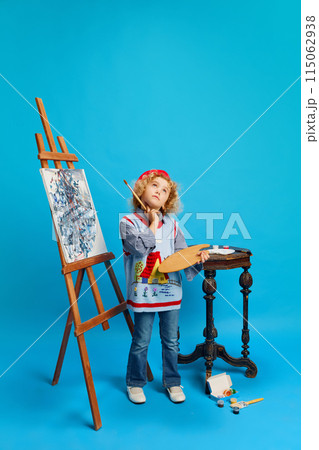 Dreaming little girl stands next to easel and creates new works of art, looking like artist against bright blue background. 115062938