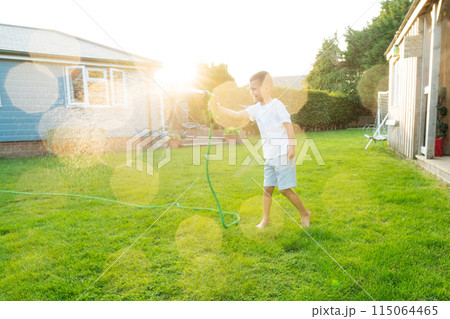 Joyful smiling boy playing with water sprinkler hose, watering grass. Summer garden outdoor fun for children. Boy splashing water on hot sunny day in backyard. Happy active childhood. Selective focus. 115064465