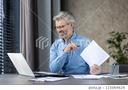 Smiling middle-aged man working at laptop home office hat home office holding papers and wearing glasses. Smiling middle-aged man working at laptop home office hat home office holding papers and wearing glasses. 115064629