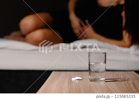 Sad young woman wearing nighty shirt suffering from menstrual pain, feeling sick to her stomach, holding belly. Girl having abdominal cramps during period and lying down on bed at home 115065235