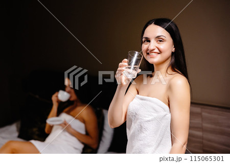 Cute young woman holding a glass of water in the morning while standing in the bedroom and behind her is her friend lying on the bed and drinking coffee Cute young woman holding a glass of water in the morning while standing in the bedroom and behind her is her friend lying on the bed and drinking coffee 115065301