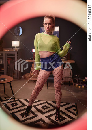 Vertical full length portrait of blonde young woman recording video at home and looking at camera through ring light Vertical full length portrait of blonde young woman recording video at home and looking at camera through ring light 115066331