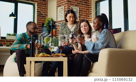 Friends at house party using smartphone to laugh at social media posts made by acquaintance while eating snacks and drinking wine. Guests using phone to look at images online, enjoying appetizers 115069132