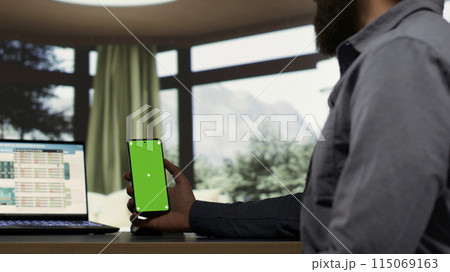Entrepreneur holding smartphone with greenscreen template while he is examining global revenue trends on laptop. Company CEO connecting on device to help employees, using isolated copyspace. 115069163