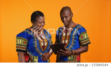 Ethnic people navigating on social media webpage, checking messages and looking at photos over orange background. African american man and woman scrolling online websites in studio. 115069164