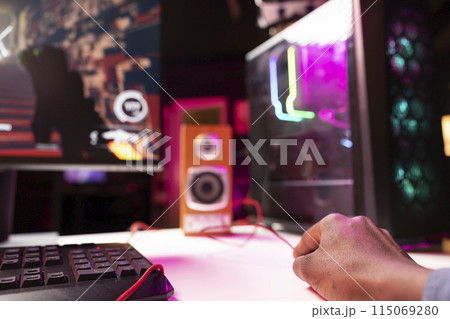 Gamer using keyboard and mouse to play FPS videogame on gaming PC tower with neon lit components. Man in apartment at night using high tech computer peripherals to defeat foes, close up 115069280