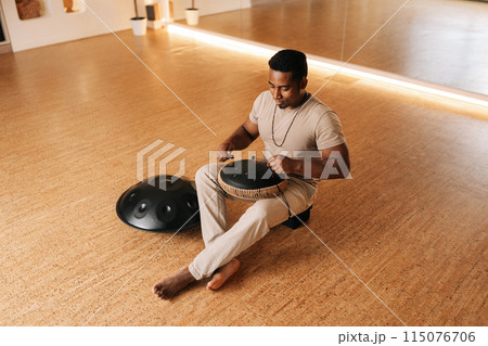 Remote top view of happy African man in trance playing meditative instrument tank drum sitting cross-legged on floor in meditation room. Concept of mindfulness, tranquility, harmony, sound therapy. Remote top view of happy African man in trance playing meditative instrument tank drum sitting cross-legged on floor in meditation room. Concept of mindfulness, tranquility, harmony, sound therapy. 115076706