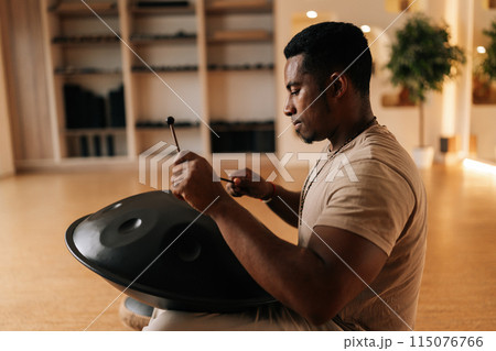 Side view of relaxed African man playing traditional ethnic music on glucophone using sticks sitting on floor in lotus pose. Concept of mindfulness, tranquility, harmony, sound therapy. 115076766