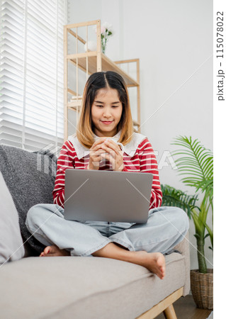 A woman is sitting on a couch with a laptop in front of her. She is holding a cup and she is working on her laptop. Concept of productivity and focus 115078022