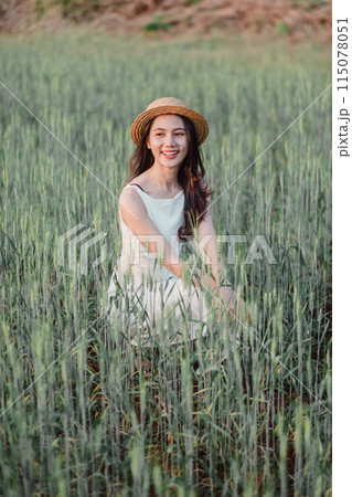 A woman is sitting in a field of tall grass, wearing a straw hat and a white dress. She is smiling and she is enjoying the peaceful surroundings 115078051