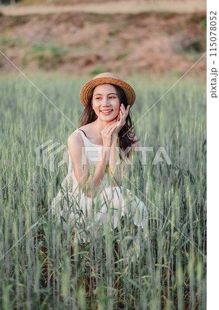 A woman is sitting in a field of tall grass, wearing a straw hat and a white dress. She is smiling and she is enjoying the outdoors 115078052