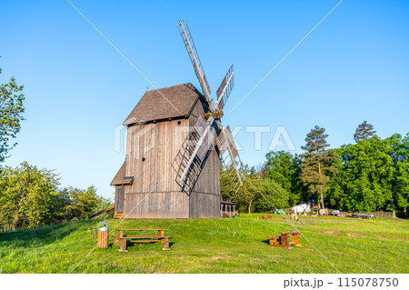 A wooden windmill stands tall against a blue sky in Borovnice, Czechia. The windmill is surrounded by lush green grass and trees. 115078750