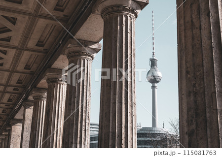 The Berlin TV Tower stands tall, framed by the classical columns of a nearby historical building on a clear day. Berlin, Germany 115081763