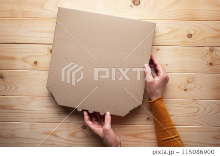 Pizza closed carton box in human caucasian hands on natural wooden table flat lay mockup with blank space Pizza closed carton box in human caucasian hands on natural wooden table flat lay mockup with blank space 115086900