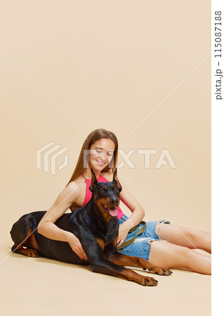 Young woman with long hair sitting on floor, smiling and hugging her large Doberman against beige studio background. 115087188