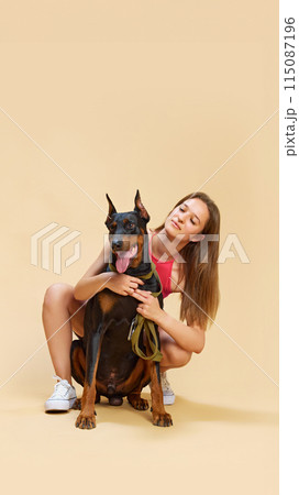 Girl with sits and hugs from behind her lovely purebred Doberman dog and cheerful smiling against beige studio background. Girl with sits and hugs from behind her lovely purebred Doberman dog and cheerful smiling against beige studio background. 115087196