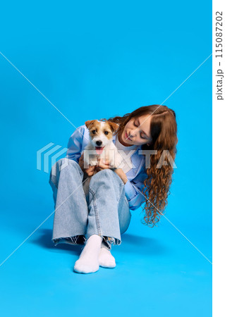 Portrait of young woman posing with her Jack Russel dog against vibrant blue studio background, enjoying time with pet. 115087202