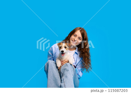 Portrait of young smiling girl in style clothes sits on floor with Jack Russel puppy against vibrant blue studio background. Portrait of young smiling girl in style clothes sits on floor with Jack Russel puppy against vibrant blue studio background. 115087203