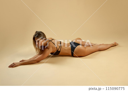 Slim woman in black lingerie lies on her stomach with her arms extended forward shows her toned back, legs against beige studio background. 115090775
