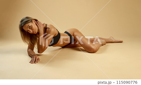 Slim and athletic woman with toned body lying elegantly on floor in black lingerie against beige studio background. 115090776