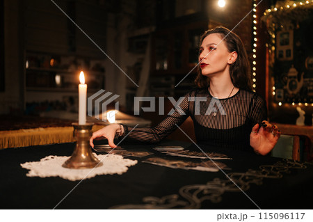 Portrait of enigmatic fortuneteller laying out tarot cards, reading signs symbols on magic cards and talks with client sitting at table in dark esoteric room with mystical atmosphere. 115096117