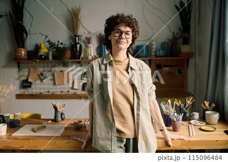 Modern female artisan wearing casual clothes with eyeglasses standing against table in workshop posing for camera 115096484
