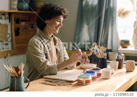 Joyful Caucasian female artisan sitting at table in workshop coloring handmade ceramic vessel with paint and paintbrush Joyful Caucasian female artisan sitting at table in workshop coloring handmade ceramic vessel with paint and paintbrush 115096510